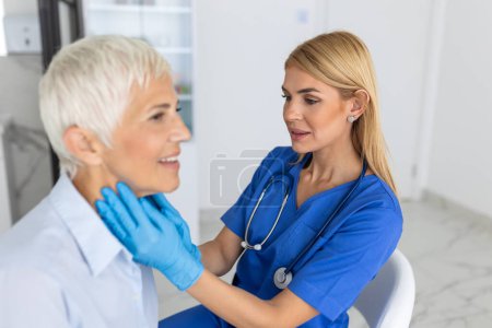 Téléchargez les photos : Femme médecin amicale portant des gants vérifiant le mal de gorge ou les glandes thyroïdes, touchant le cou de la patiente âgée visitant le bureau de la clinique. Concept de prévention du cancer de la thyroïde - en image libre de droit