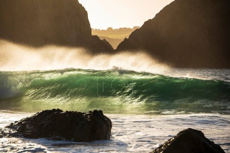 Foto de Imagen épica del paisaje de olas turquesas de jade que se estrellan en la orilla y rocas en Kynance Cove Cornwall con resplandeciente fondo de salida del sol y gotas de rocío de agua en el viento - Imagen libre de derechos