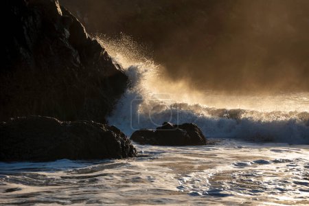 Photo for Epic landscape image of jade turquoise waves crashing onto shore and rocks in Kynance Cove Cornwall with glowing sunrise background and water spray droplets in wind - Royalty Free Image