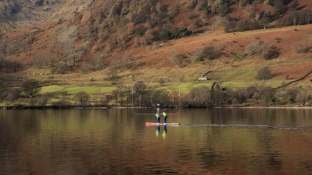 Téléchargez les photos : Beautiful Winter landscape views of mountain ranges around Ullswater in Lake District viewed from boat on lake with unidentified people paddle boarding on lake - en image libre de droit