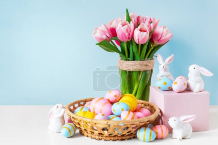 Photo for Easter and spring holiday greeting card concept. Pink tulips, easter bunnies with colorful eggs in basket and blue background. Painted Easter eggs in nest and tulips with white bunny. - Royalty Free Image
