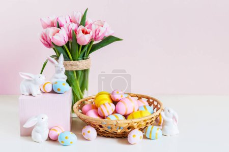 Photo for Easter and spring holiday greeting card concept. Pink tulips, easter bunnies with colorful eggs in basket. Painted Easter eggs in nest and tulips with white bunny, space for text. Copy space. - Royalty Free Image