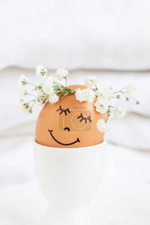 Photo for Easter egg with cute face in floral wreath crowns in egg cup on white background. Easter egg with flowers and sleepy eyes in sunny light. Happy Easter concept. - Royalty Free Image