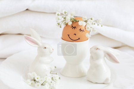 Photo for Easter egg with cute face in floral wreath crowns in egg cup with two white easter bunnies. Easter egg with flowers and sleepy eyes in sunny light and beautiful bunnies. - Royalty Free Image