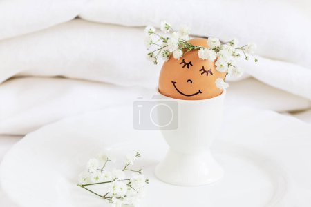 Photo for Easter egg with cute face in floral wreath crowns in egg cup on white background. Easter egg with flowers and sleepy eyes in sunny light. Happy Easter concept. - Royalty Free Image