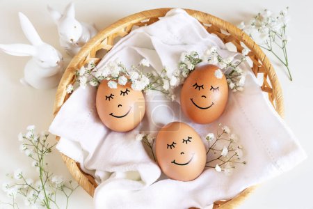 Photo for Three happy Easter eggs with cute faces in floral wreath crowns on white background in nest and two white bunnies. Easter eggs with flowers and sleepy eyes in sunny light with cute two white bunnies. - Royalty Free Image