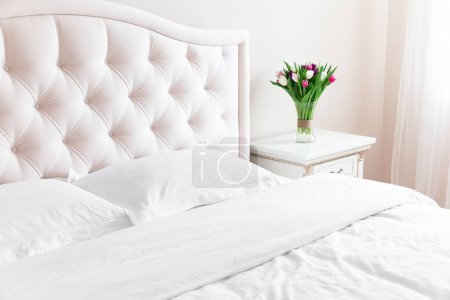 Photo for Bedroom with bed, white bedding, and bedside table with bouquet of tulips in a vase. White pillows, duvet and duvet case on bed with beige headboard.  Bed with clean white pillows and bed sheets. - Royalty Free Image