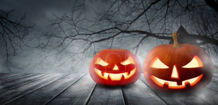 Photo for Two spooky Halloween pumpkins, Jack O Lantern, with an evil faces and eyes on a wooden table with a misty gray background. Halloween Pumpkins with dark background. - Royalty Free Image