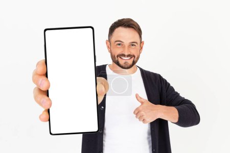 Foto de Joven guapo sosteniendo una maqueta de smartphone con una pantalla en blanco y sonriendo sobre un fondo blanco. Celebrando el éxito. Gadget con espacio vacío para maqueta, banner, aplicación. - Imagen libre de derechos