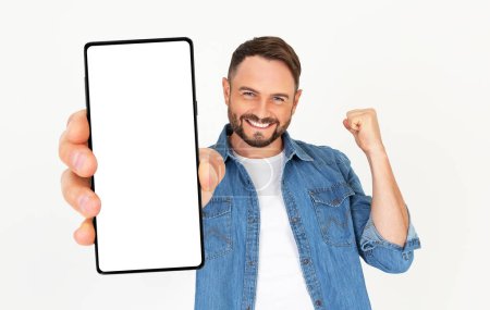 Foto de Joven guapo sosteniendo una maqueta de smartphone con una pantalla en blanco y sonriendo sobre un fondo blanco. Celebrando el éxito. Gadget con espacio vacío para maqueta, banner, aplicación. - Imagen libre de derechos