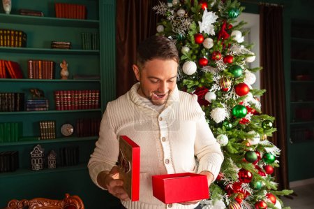 Foto de Joven excitado abriendo regalo de Navidad en su casa. Retrato de feliz hermoso hombre emocional apertura de la caja de regalo de Navidad. - Imagen libre de derechos