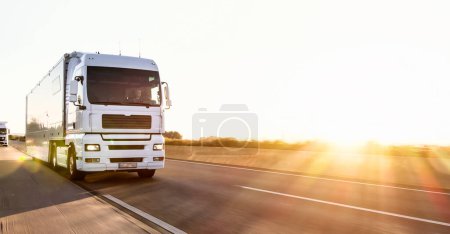 Photo for Cargo truck with cargo trailer driving on a highway. White Truck delivers goods in early hours of the Morning - very low angle drive thru close up shot. - Royalty Free Image
