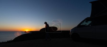 Photo for Surferboy sitting near his mini van and looking on the ocean at summer sunset  with a surfboard on her side - Royalty Free Image