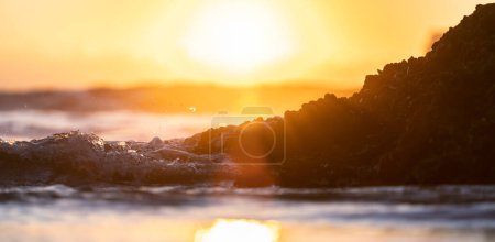 Photo for Close up shot of waves hitting a stone at the beach at calm orange sunset ocean. selective focus shot - Royalty Free Image