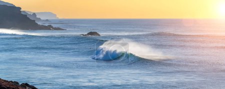 Téléchargez les photos : Énorme belle vague se brise à la côte tandis qu'une brise souffle l'eau de crachat hors de la mer à un coucher de soleil merveilleux avec ciel orange - en image libre de droit