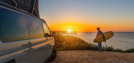 Téléchargez les photos : Surferboy sitting near his mini van and looking on the ocean at summer sunset  with a surfboard on her side - en image libre de droit