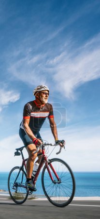 Photo for Mature Adult on a racing bike climbing the hill at mediterranean sea landscape coastal road - vertical shot for your story, reel - Royalty Free Image