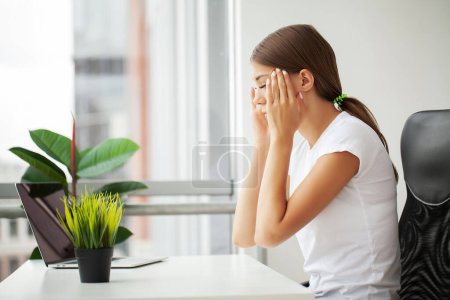 Schöne gestresste junge Büroangestellte sitzt am Schreibtisch und hält den Kopf wegen Schmerzen im Büro.