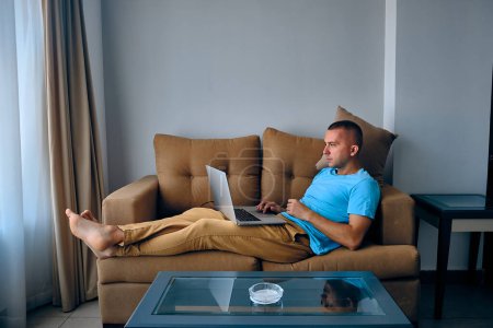 Téléchargez les photos : Jeune homme caucasien adulte travaille sur un ordinateur portable couché sur un canapé. Homme réfléchi surfant sur un filet couché sur le canapé - en image libre de droit
