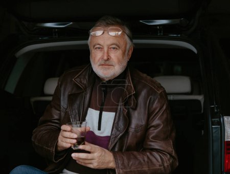 Foto de Anciano con una barba gris en una chaqueta de cuero sostiene en su mano un vaso de whisky sobre el fondo de un tronco abierto de una ca - Imagen libre de derechos