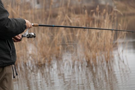 Photo for The hands of a spinner turn the handle of a spinning reel on a telescopic fishing rod on an autumn day. Blurred dry reeds in the backgroun - Royalty Free Image