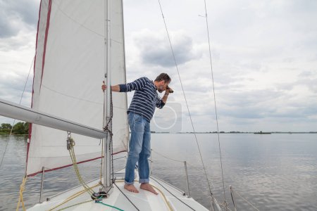 Photo for Captain holds on to the mast. A man in a striped sweater and jeans sits on the nose of a sailing yacht and looks through binocular - Royalty Free Image
