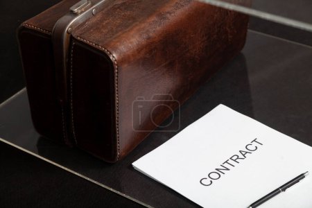 Photo for Business attributes. Close-up of a contract printed on paper and a leather carpetbag in a transparent box. The pen is on top of the contrac - Royalty Free Image