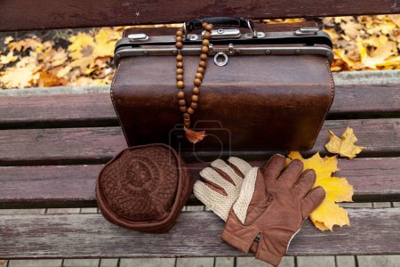 Photo for Still life with a valise and a rosary. There is a leather valise on a wooden bench in the park. There is a wooden rosary on it. Nearby lies a hat and glove - Royalty Free Image