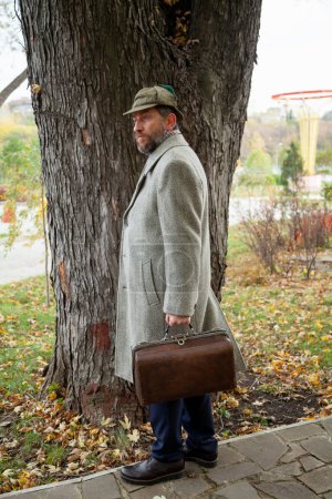Photo for Waiting in the autumn park. A man in a gray coat stands in front of the trunk of a large tree in the park. Holding a carpetbag in his han - Royalty Free Image