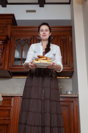 Photo for Cute brunette woman in vintage clothes holds a plate with delicious pies in a wooden kitchen. Bottom vie - Royalty Free Image