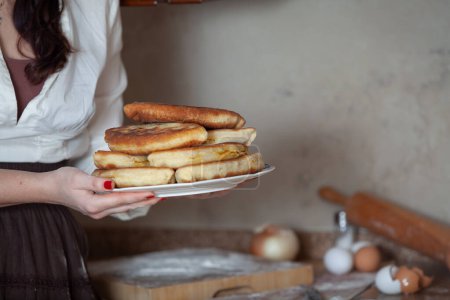 Photo for A woman holds a plate with fried pies, in the background there is a table, eggs, an onion, a rolling pi - Royalty Free Image
