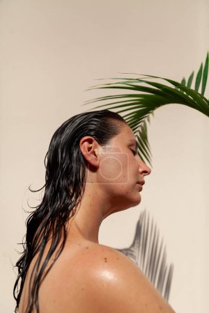 Photo for A woman with a bare back and wet dark hair stands in profile under a green palm branch. On a light background, a graceful shadow from a palm tre - Royalty Free Image