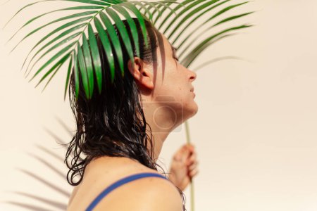 Photo for Girl and palm branch. A beautiful girl in a swimsuit holds in her hands a branch of the areca palm tree on a light background. Palm tree provides interesting shad - Royalty Free Image