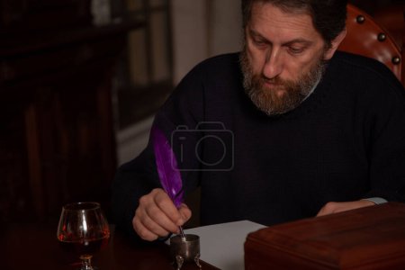 Un homme barbu s'assoit à une grande table polie et écrit sur une feuille blanche avec un stylo plume et une encrier. Il y a un verre avec un patin devant salut