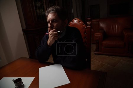 A man with a beard sits at a large polished table and ponders over a white sheet. In his hand is a wooden pen with a metal nib. There is a metal inkwell nearb