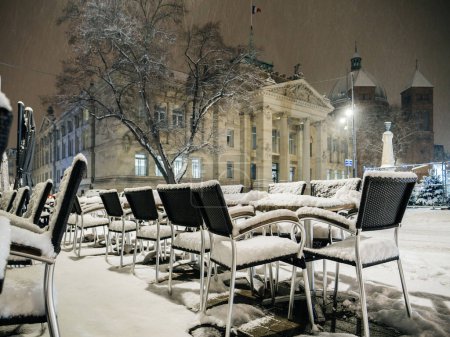Téléchargez les photos : Vue d'un restaurant avec chaises et table recouvertes de neige par une froide nuit d'hiver avec magnifique palais haussmannien en arrière-plan - en image libre de droit