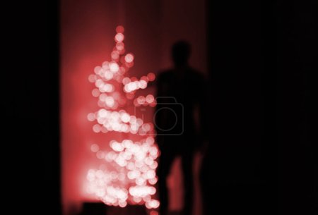 Téléchargez les photos : Male silhouette staying near a defocused red Christmas fir tree - concept for loneliness during winter holidays, consumerism etc - en image libre de droit
