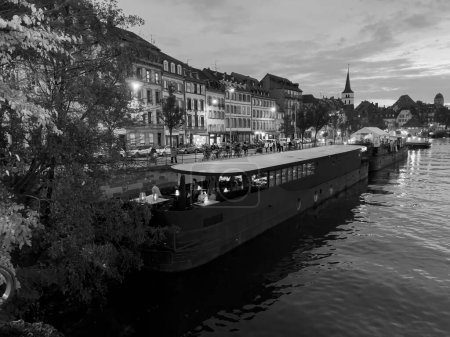 Téléchargez les photos : Strasbourg, France - Oct 29, 2022: Monochrome scene Strasbourg at dusk with restaurant boats full with people celebrating pre-Halloween parties - calm waters of Ill river - en image libre de droit