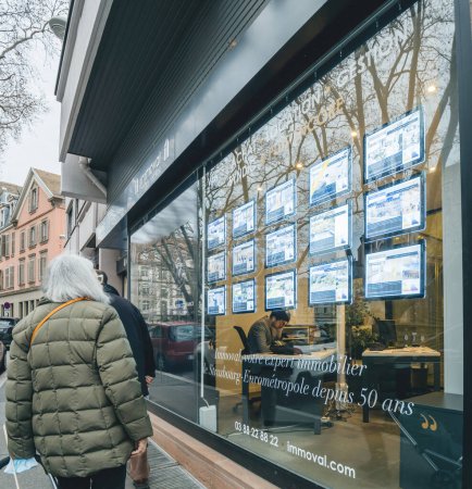 Téléchargez les photos : Strasbourg, France - Jan 5 2023: Senior woman looking at the prices of houses and apartments at a real estate agency glass showcase window with multiple announcement - en image libre de droit