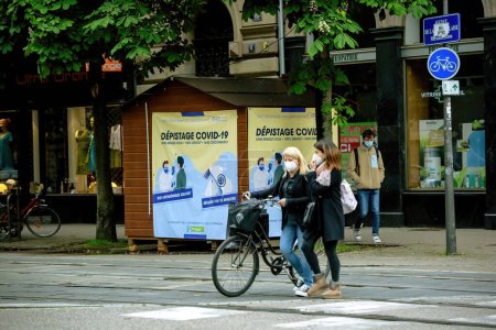 Foto de Strasbourg, France - May 20, 2021: Two female friends crossing street with Depistage Covid translated as Covid Screening booth in background - Imagen libre de derechos