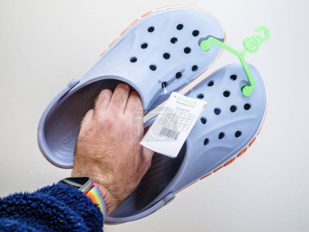 Photo for Paris, France - Dec 2, 2022: POV male hand holding pair of new Crocs sandals foam clogs in blue color with red inscription - Royalty Free Image