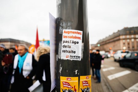 Foto de Strasbourg, France - Jan 19, 2023: Rise the wages sticker on street pillar - defocused people at protest against the French governments planned pension reform to push the retirement age from 62 to 64 - Imagen libre de derechos