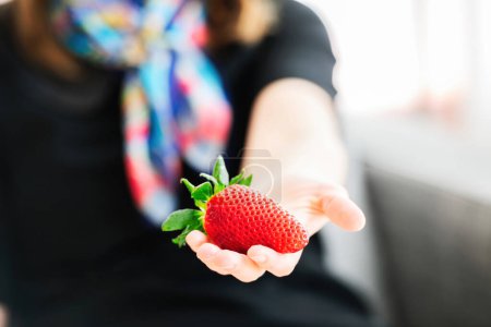 Foto de Female hand holding single strawberry - silhouette of woman in background with colored scarf - Imagen libre de derechos