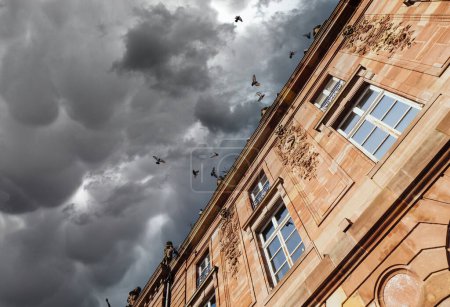 Téléchargez les photos : Dramatic storm clouds sky low angle view of stone facade decorated with diverse romantic bas relief and large crowd of pigeons flying on the blue sky background - en image libre de droit