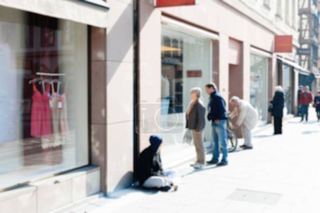 Foto de Biew desenfocado desenfoque de mendigo en la calle de una ciudad moderna con silueta de pareja admirando junto a él productos en un escaparate - Imagen libre de derechos