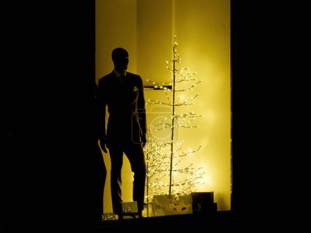 Téléchargez les photos : Vitrine avec costume masculin couture de luxe à côté d'un arbre de Noël illuminé abstrait - tons chauds jaunes la nuit - en image libre de droit