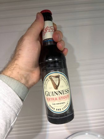 Foto de Alemania, Múnich - Apr 13, 2022: POV mano masculina sosteniendo la botella de cerveza Guinness Extra Stout - fondo de cocina - Imagen libre de derechos