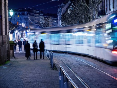 Foto de Una escena nocturna de la ciudad con un grupo de personas iluminadas por el tren que pasa, creando un movimiento borroso en una zona urbana llena de estructuras construidas y varios modos de transporte. - Imagen libre de derechos