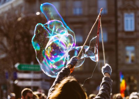 Foto de Una mujer está usando una varita para hacer burbujas líquidas en un festival irreconocible, trayendo alegría y diversión para la multitud mientras observan en anticipación de estas frágiles creaciones en el aire. - Imagen libre de derechos
