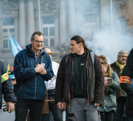 Téléchargez les photos : Strasbourg, France - 29 mars 2023 : Une foule d'adultes, pour la plupart des hommes en jeans et vestes en signe de protestation avec de la fumée en arrière-plan - La France connaît des semaines de protestations et de grèves liées à une hausse des - en image libre de droit